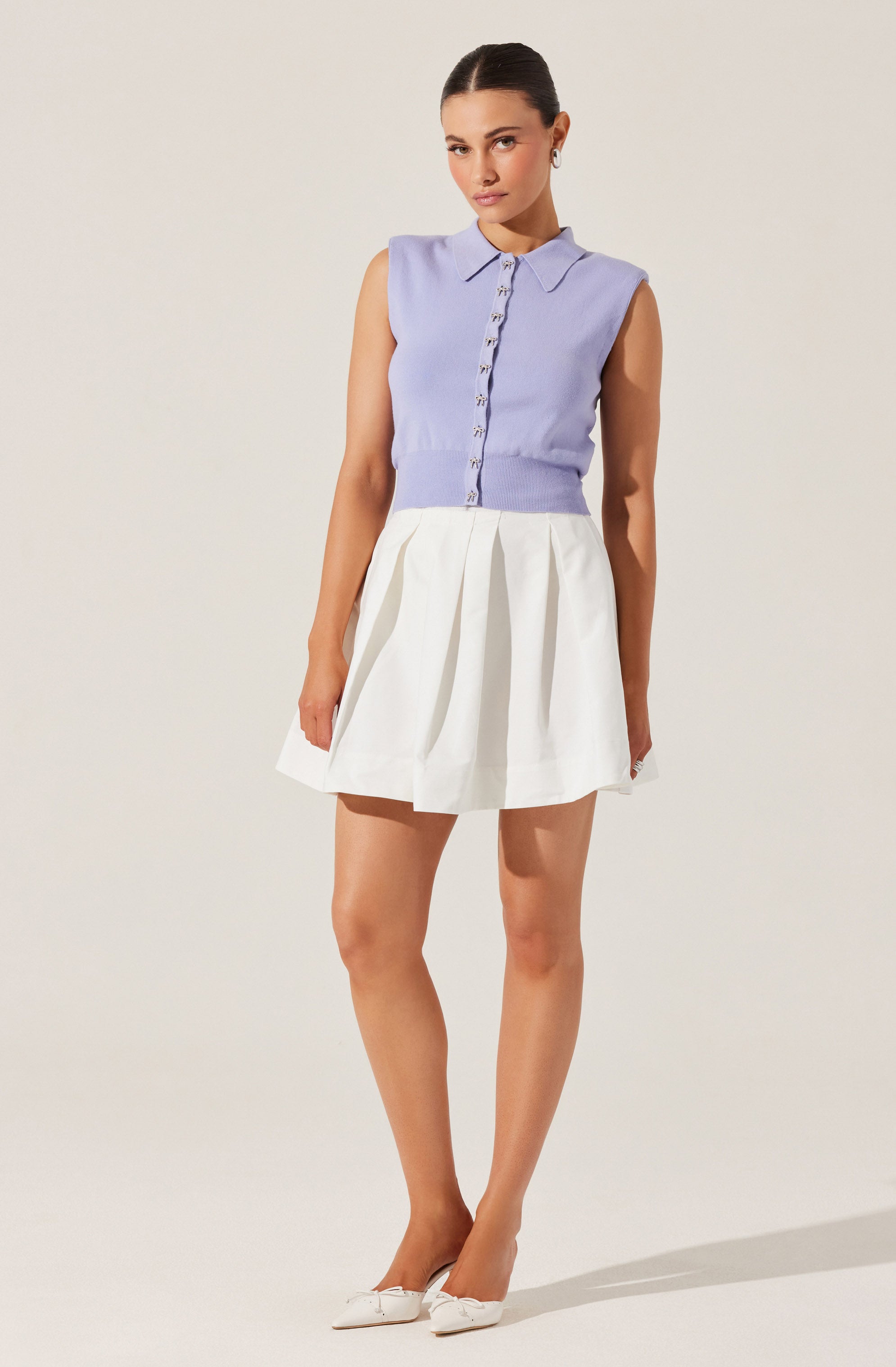 The outfit consists of a sleeveless, lavender-colored button-up top with a collared neckline, paired with a high-waisted, white, pleated skirt that falls above the knees. The woman is also wearing white shoes with a small bow detail.