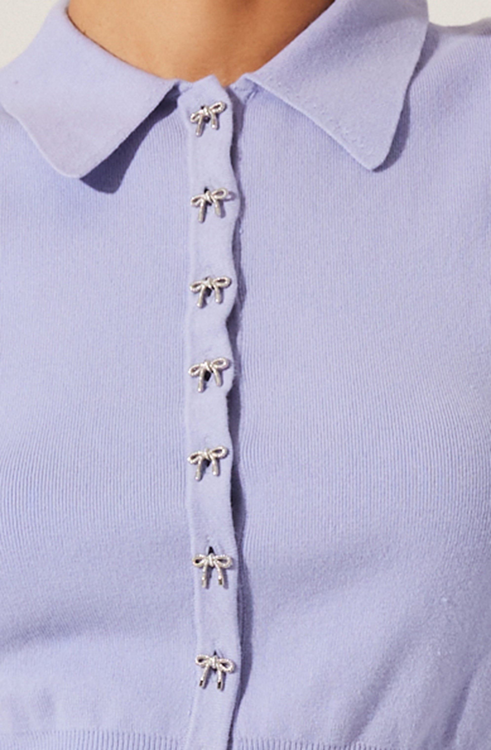 A close-up of a light purple collared shirt featuring small decorative bows along the button placket, which are metallic and shiny.