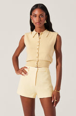 A woman is wearing a sleeveless, cream-colored button-up top with a collar. She pairs it with matching high-waisted shorts that have a textured fabric. She accessorizes with gold earrings, a bracelet, and a ring.