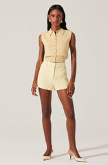A woman is wearing a sleeveless, light beige button-up top with a collar, tucked into matching high-waisted, tailored shorts. She is accessorized with gold earrings, a gold bracelet, and rings. She has on white pointed-toe high heels with a small heel.