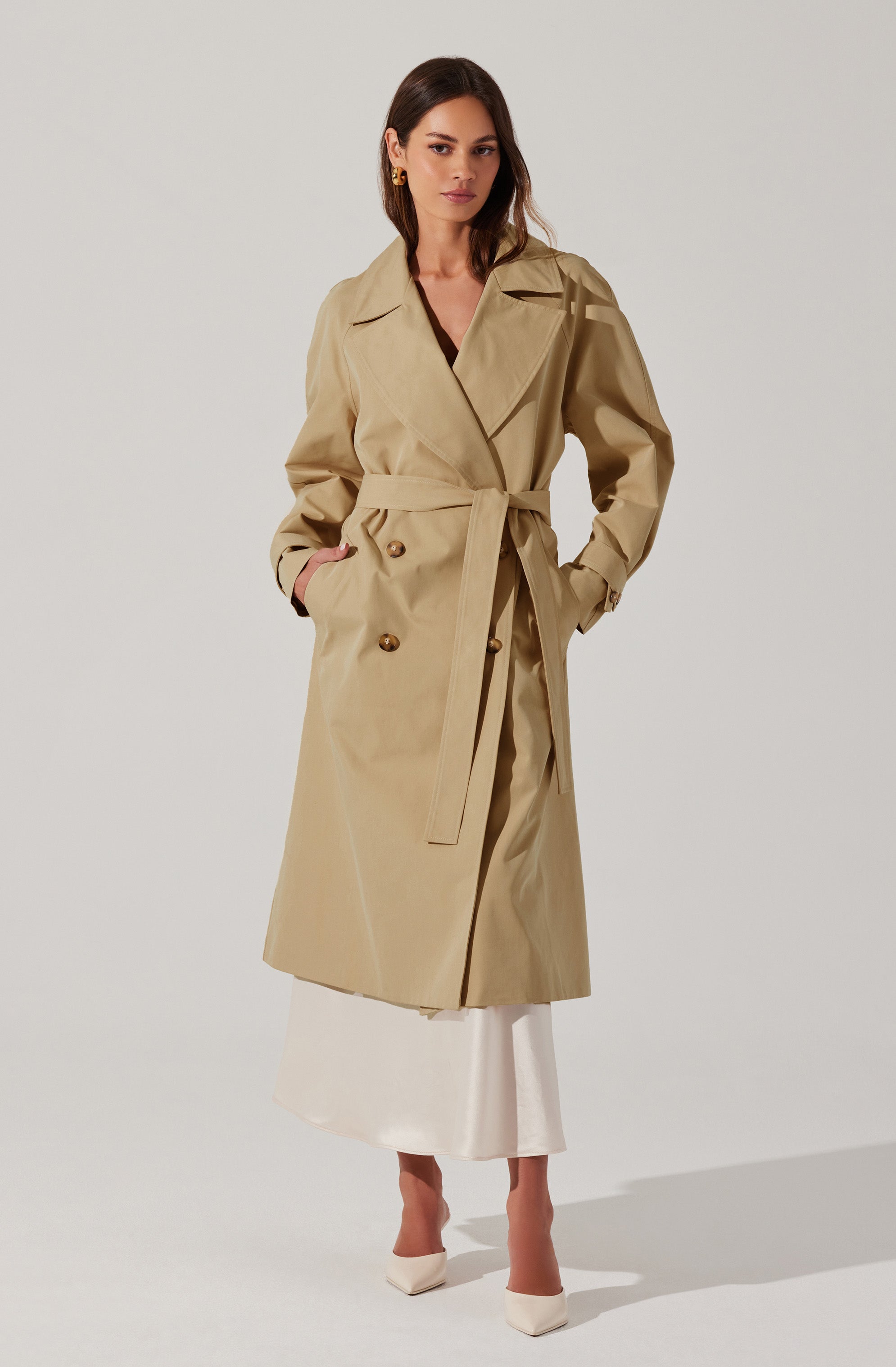 A woman is wearing a classic beige trench coat with a belted waist, double-breasted buttons, and a wide lapel. The coat has long sleeves and side pockets. She pairs it with a white skirt and white high-heeled shoes. She also has small gold earrings.
