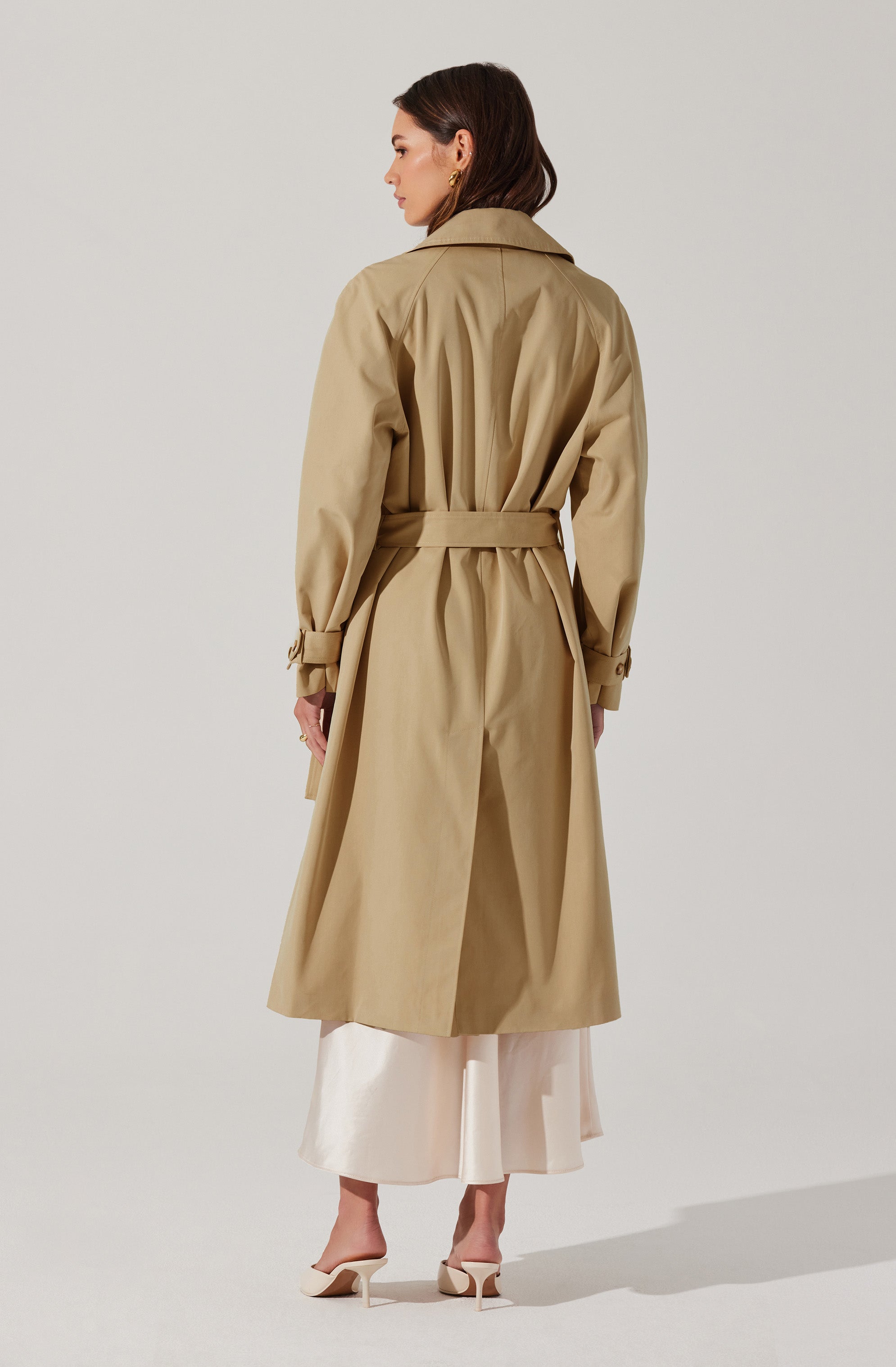 A woman is wearing a long beige trench coat with a belt at the waist, buttoned cuffs on the sleeves, and a classic collar. She has paired the coat with a light-colored, ankle-length dress and beige high-heeled shoes. The coat's design includes pleats and a vent at the back.