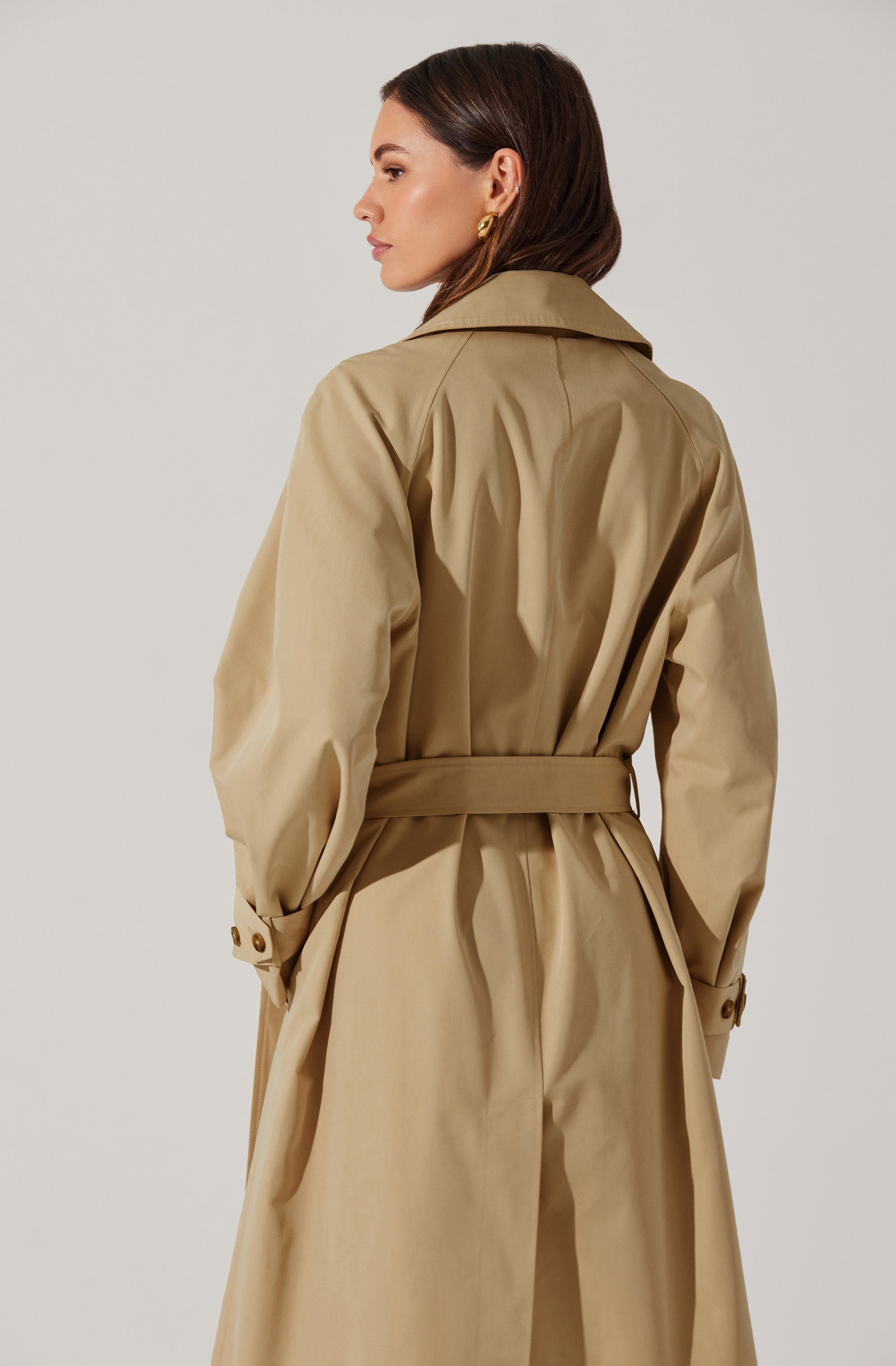 A woman is wearing a beige trench coat with a wide collar, long sleeves with buttoned straps at the cuffs, and a matching belt cinched at the waist. The coat features a classic design with a smooth, slightly shiny fabric.