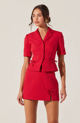 A woman is wearing a coordinated red outfit consisting of a tailored short-sleeve blazer with a notched collar and front button closure, and a matching high-waisted mini skirt with a front slit and decorative bow ties on the sides and front. The blazer features a fitted silhouette with darts at the waist, and the skirt is form-fitting. She has long, dark hair styled in loose waves and is accessorized with a pair of gold earrings.