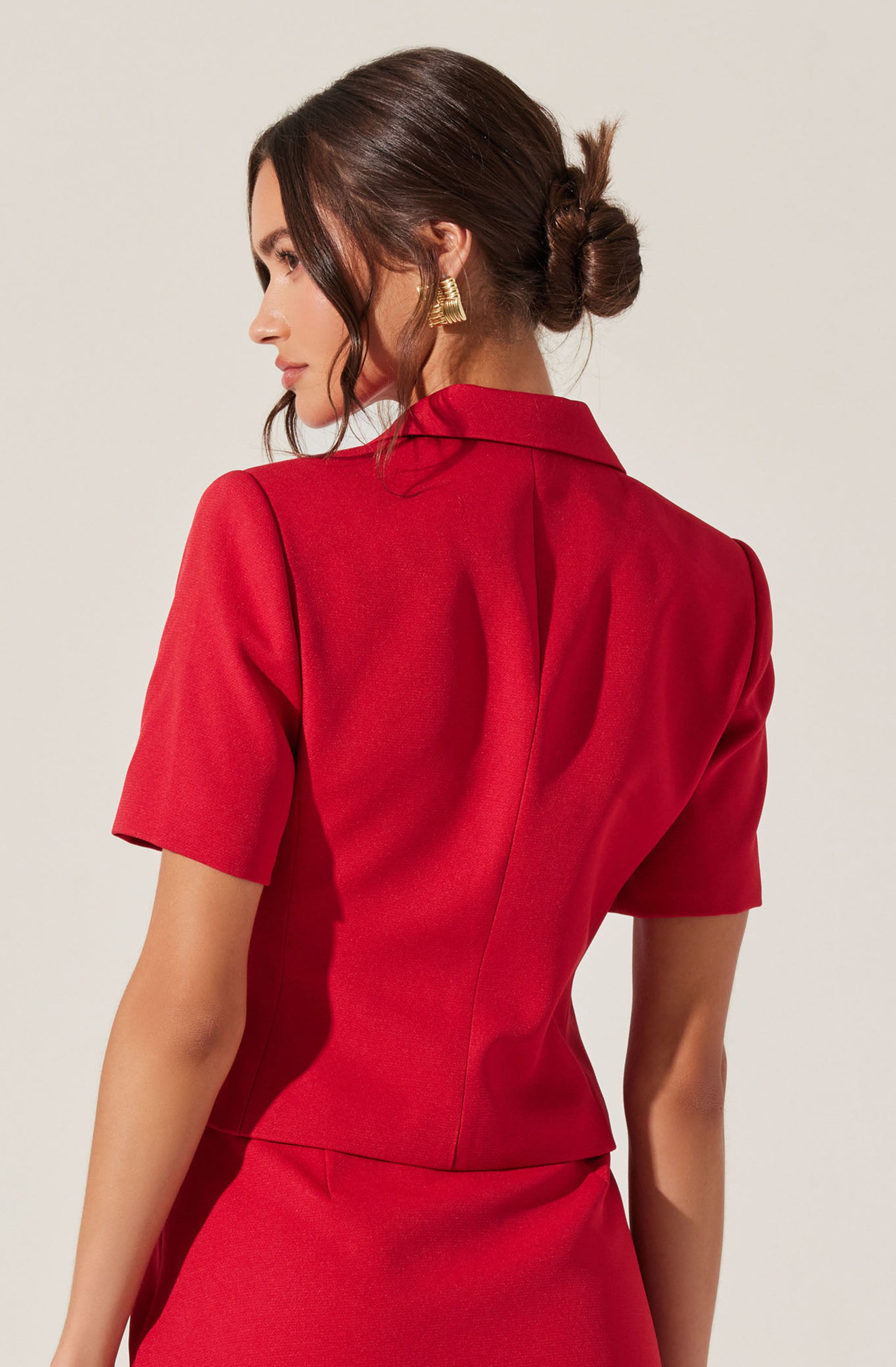 A woman is wearing a red short-sleeved blazer with a structured collar and a fitted waist. She also has on matching red skirt and is accessorized with gold statement earrings. Her dark brown hair is styled in a low, messy bun with loose strands framing her face.
