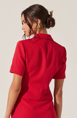 A woman is wearing a red short-sleeved blazer with a structured collar and a fitted waist. She also has on matching red skirt and is accessorized with gold statement earrings. Her dark brown hair is styled in a low, messy bun with loose strands framing her face.