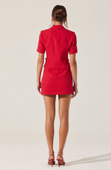 A woman is wearing a matching red short-sleeved blazer and a red mini skirt. The blazer is tailored with a structured fit, and the skirt is fitted and straight. She is also wearing red high-heeled shoes with ankle straps. The outfit has a sleek and polished appearance.