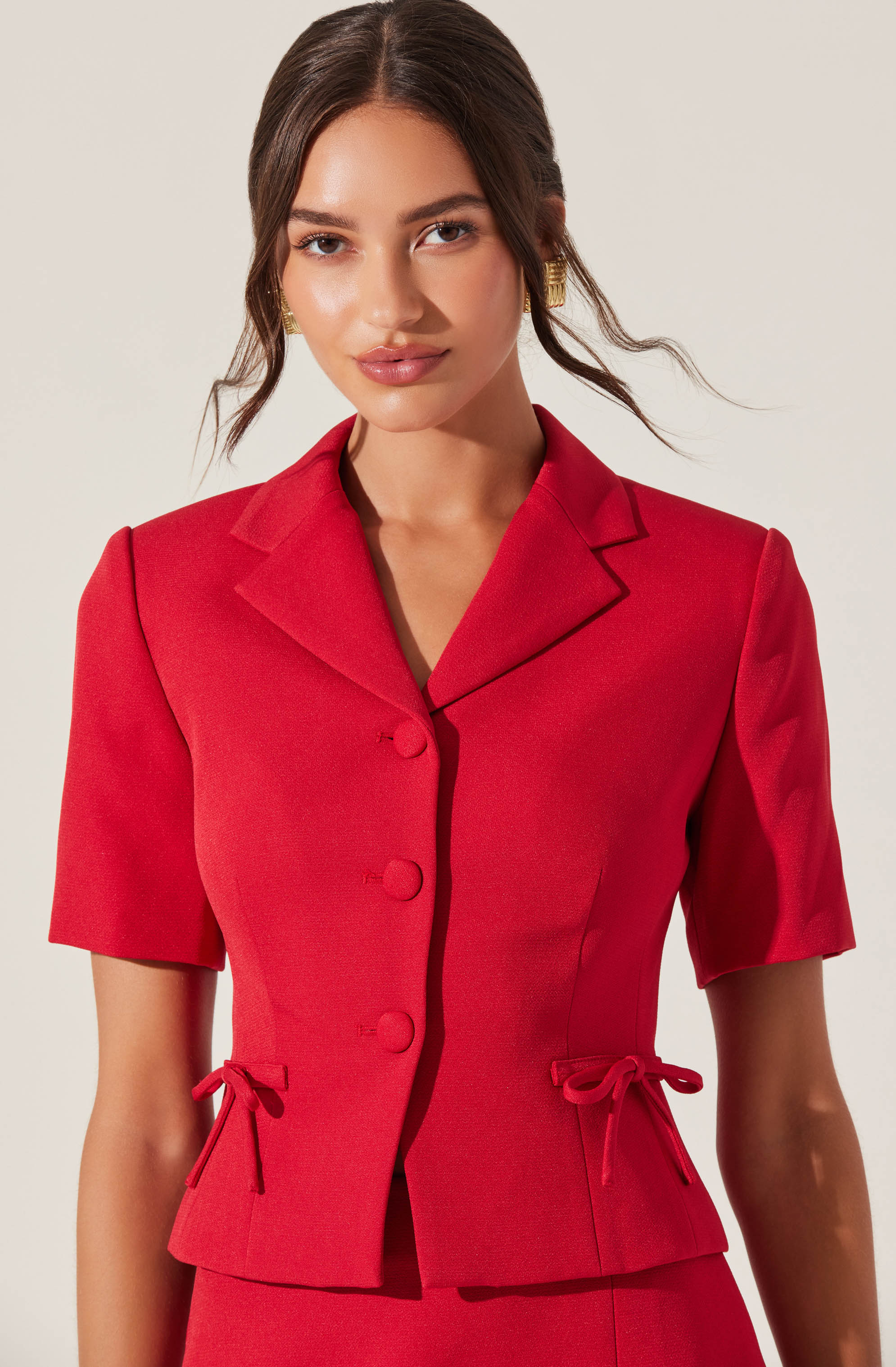 A woman is wearing a fitted, short-sleeved red blazer with a notched lapel collar and three matching buttons down the front. The blazer features decorative ties at the waist on both sides.