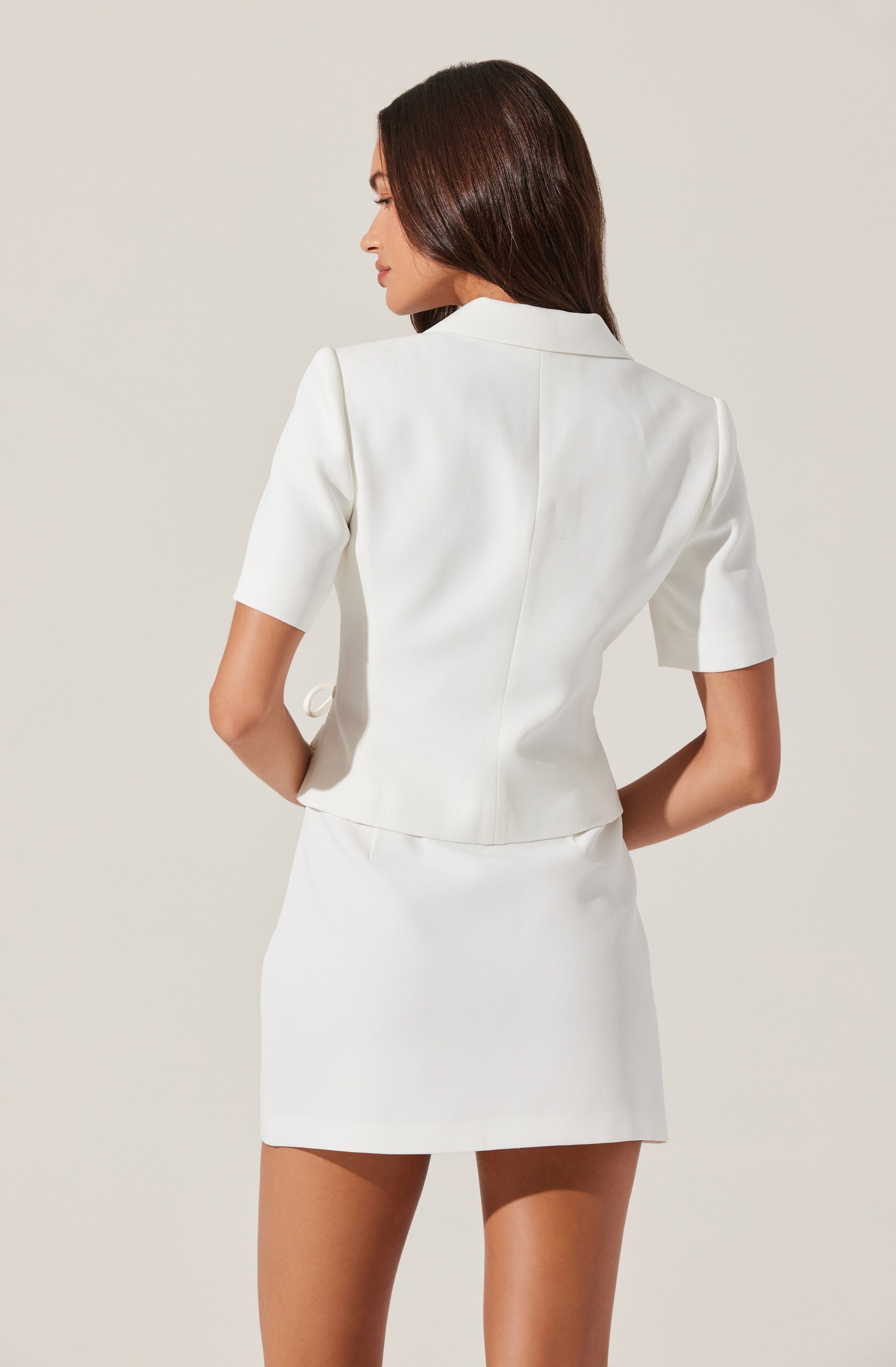 A woman is wearing a white, short-sleeved blazer and a matching white skirt. The blazer has a structured fit with a collar and front seams, while the skirt is a straight, knee-length design. The outfit is clean, minimalist, and elegant.