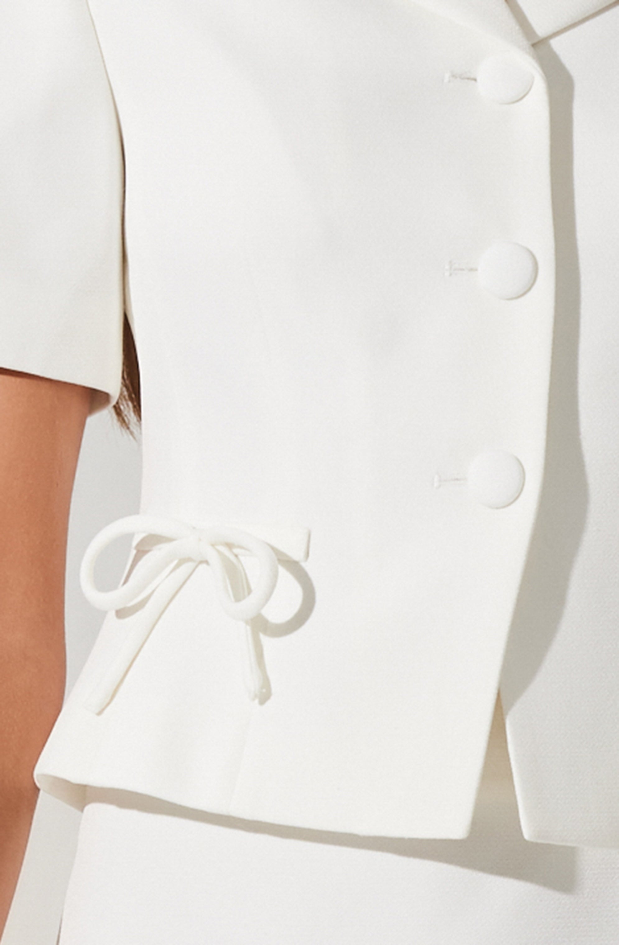 A woman is wearing a white, structured garment with large, round buttons down the front and a tied bow detail on the side near her waist. The fabric appears to be thick and smooth, giving a tailored look. The garment covers her torso and part of her arm.