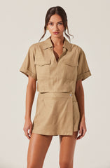 A woman is wearing a beige short-sleeved cropped button-up shirt with a collar and two flap pockets on the chest. She also has on a high-waisted beige skirt with a front button closure.