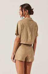 A woman is wearing a beige, short-sleeved, cropped jacket with rolled cuffs and a collar, paired with matching high-waisted shorts that have visible back pockets.
