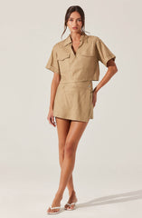 A woman is wearing a matching two-piece outfit in a beige color. The top is a short-sleeved, collared shirt with flap pockets on the chest. The skirt is a high-waisted, A-line mini skirt with a slight slit on one side. She is also wearing a pair of white high-heeled sandals.