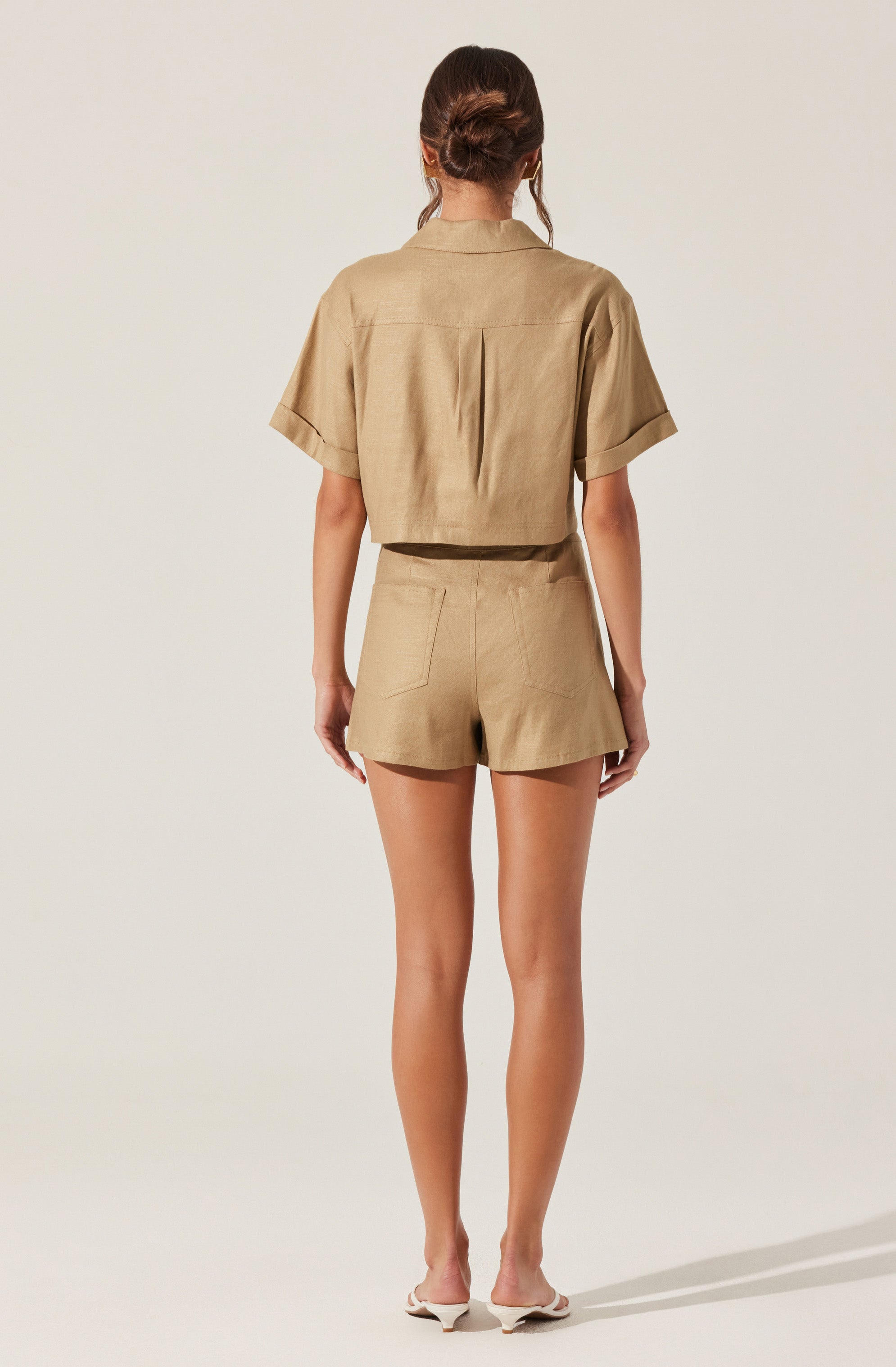 A woman is wearing a beige short-sleeved button-up shirt with a collar and a back yoke, paired with matching high-waisted shorts that have a tailored fit and a single back pocket. She has her hair styled in a loose bun and is standing against a plain, light-colored background.