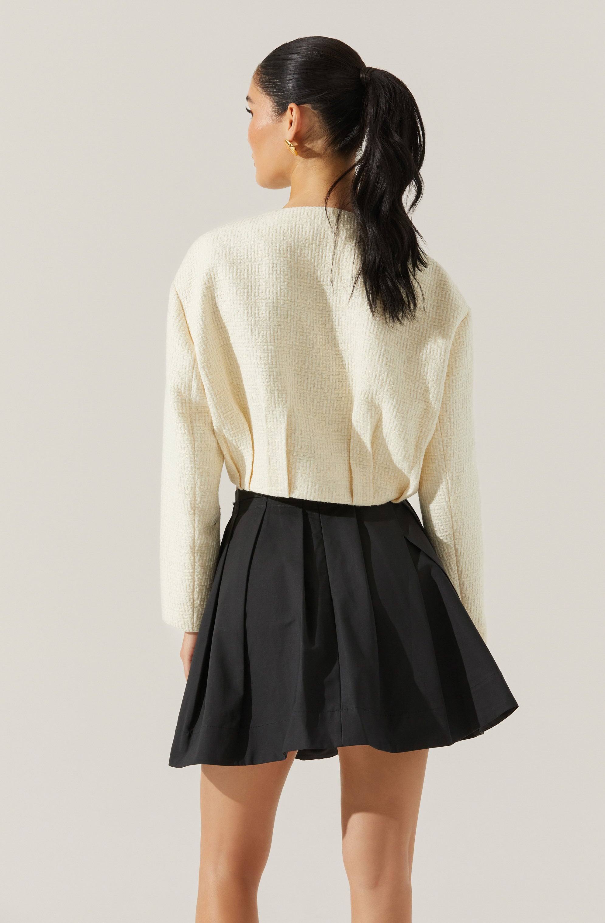 A woman is wearing an oversized, cream-colored knit sweater with a textured pattern, paired with a high-waisted, black pleated skirt that has a slightly flared silhouette. Her dark hair is styled in a high ponytail.