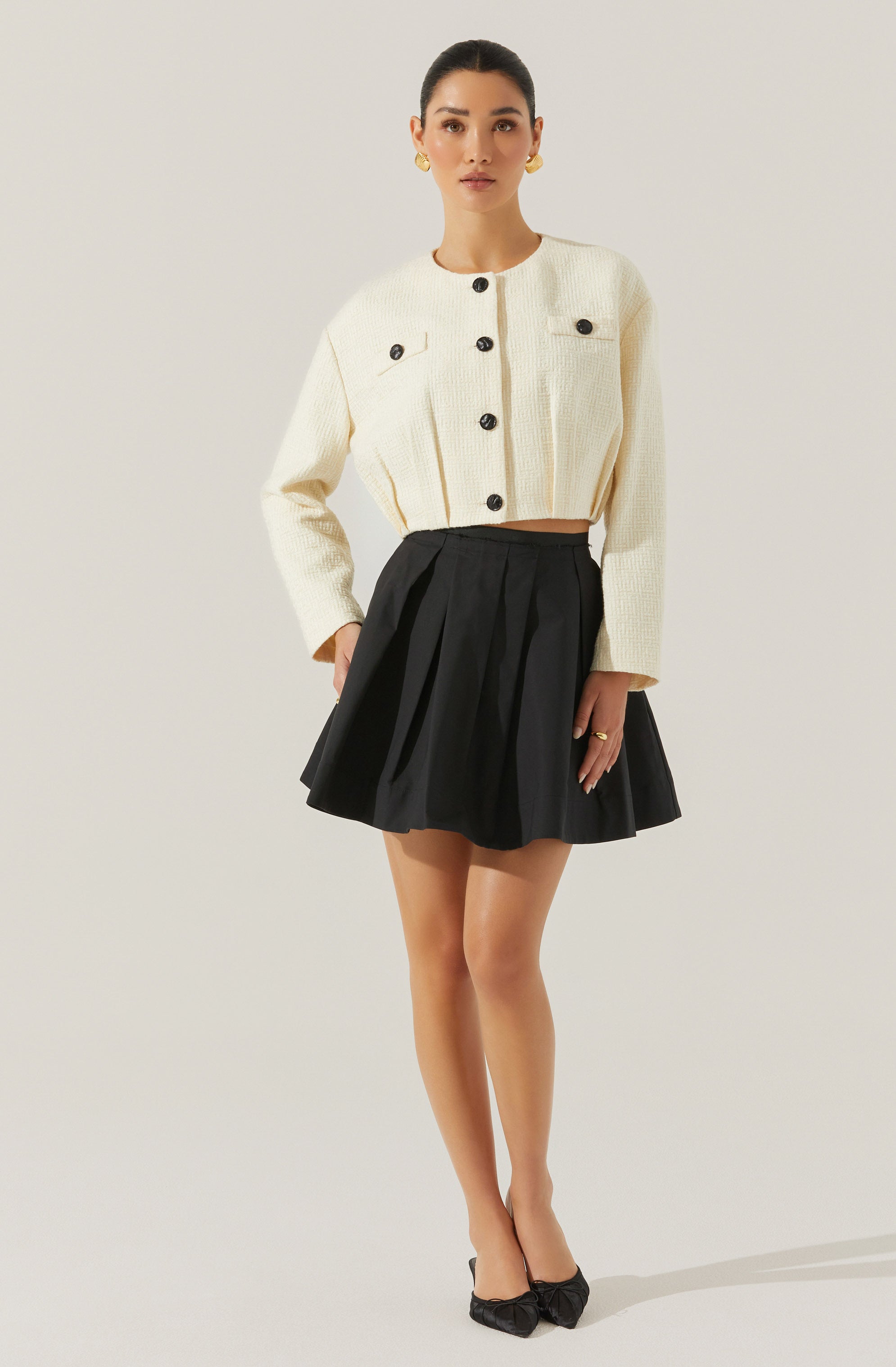 A woman is wearing a cream-colored, textured, long-sleeve cropped jacket with large black buttons, paired with a high-waisted black pleated skirt. She accessorizes with gold earrings and rings, and is wearing black pointed-toe heels with bows.