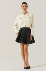 A woman is wearing a cream-colored, textured, long-sleeve cropped jacket with large black buttons, paired with a high-waisted black pleated skirt. She accessorizes with gold earrings and rings, and is wearing black pointed-toe heels with bows.