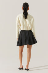 A woman is wearing a cream-colored, loose-knit sweater with long sleeves, paired with a high-waisted, pleated black skirt that reaches mid-thigh. She has on black open-toe mules with small heels and is accessorized with large round earrings.