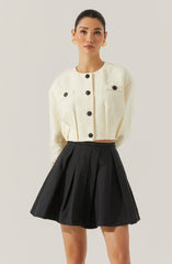 A woman is wearing a cream-colored, textured, cropped cardigan with large black buttons and flap pockets on the chest. She pairs it with a high-waisted black pleated skirt. She accessorizes with gold earrings.