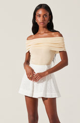 A woman is wearing an off-the-shoulder, cream-colored top with gathered detailing across the chest and sleeves. She pairs it with high-waisted white shorts that have a subtle, eyelet pattern.