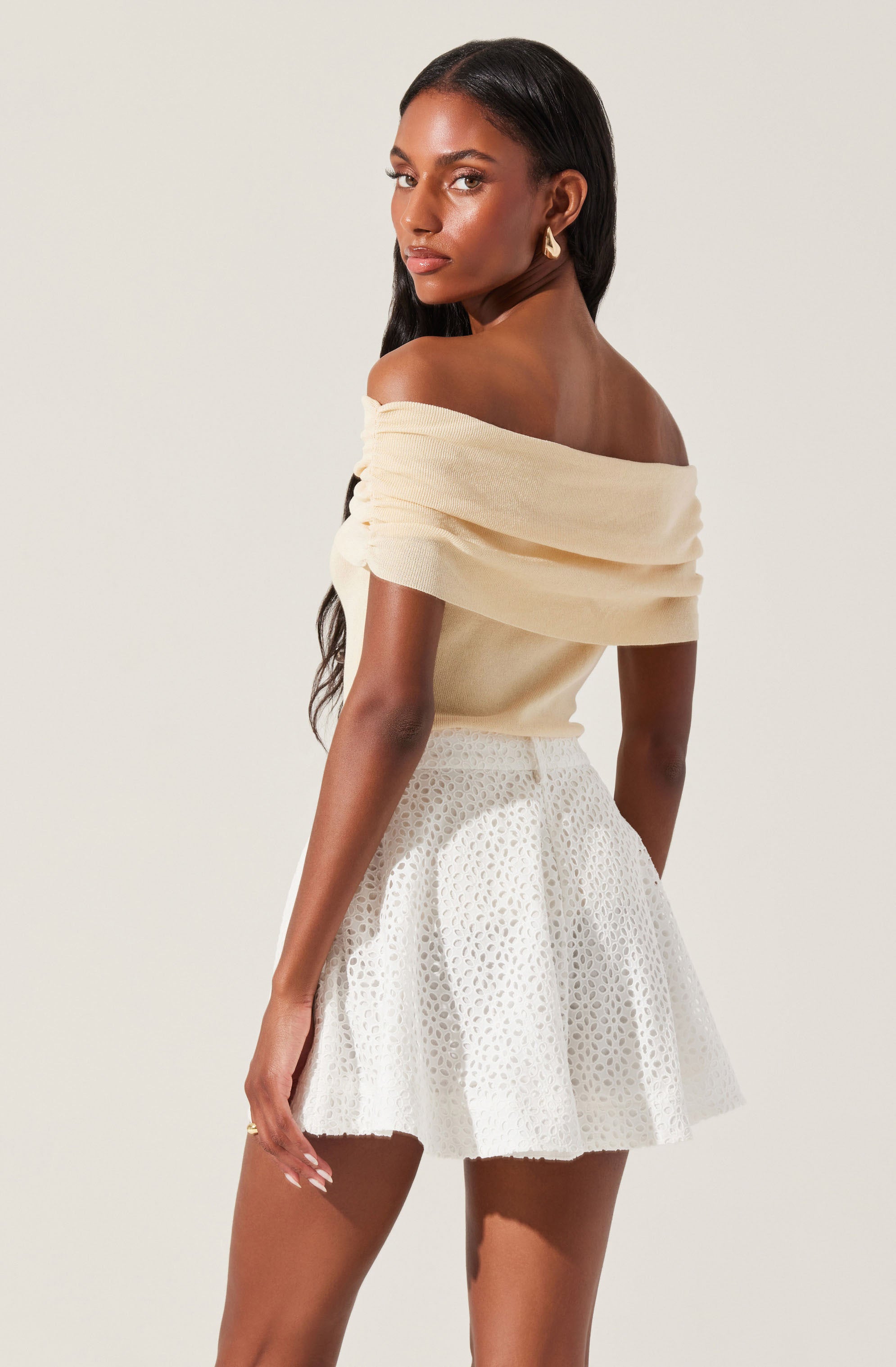 A woman is wearing an off-the-shoulder, cream-colored top with soft, draped fabric. She pairs it with a white, high-waisted skirt featuring a textured, eyelet pattern. She accessorizes with gold hoop earrings and has long, dark hair styled in loose waves.