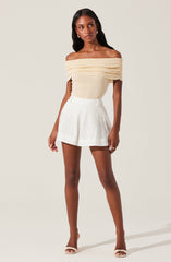 A woman is wearing an off-the-shoulder, cream-colored top with layered, ruched fabric around the neckline. She pairs it with high-waisted white shorts that have a subtle eyelet pattern. She completes the look with nude high-heeled sandals.