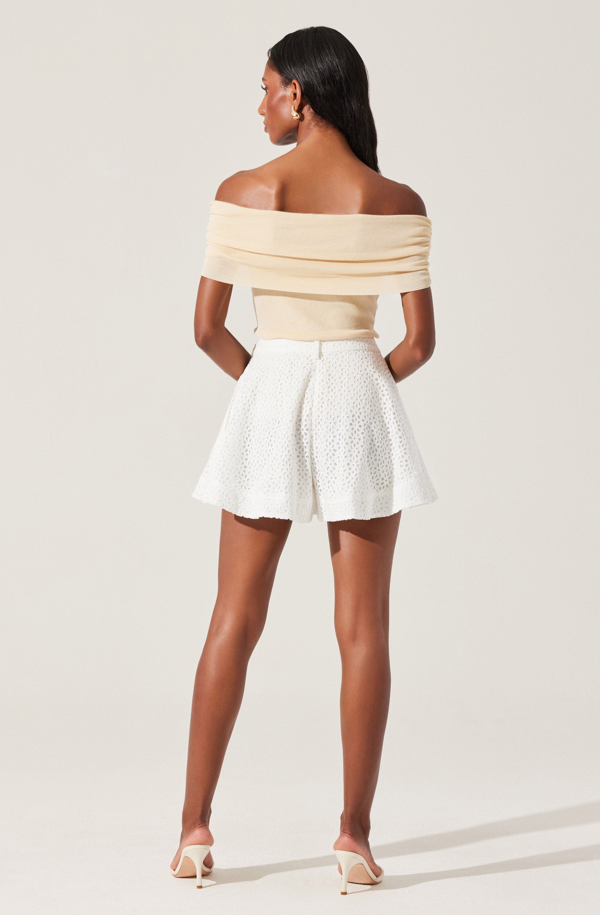 A woman is wearing an off-the-shoulder cream-colored top with wide ruffled bands around the shoulders and upper arms. She pairs it with high-waisted white shorts that have a textured, eyelet pattern and a flared, A-line silhouette. She is also wearing light-colored high-heeled shoes.
