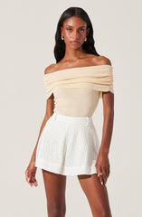 A woman is wearing an off-the-shoulder beige top with gathered fabric across the neckline and white high-waisted shorts with a textured, eyelet pattern.