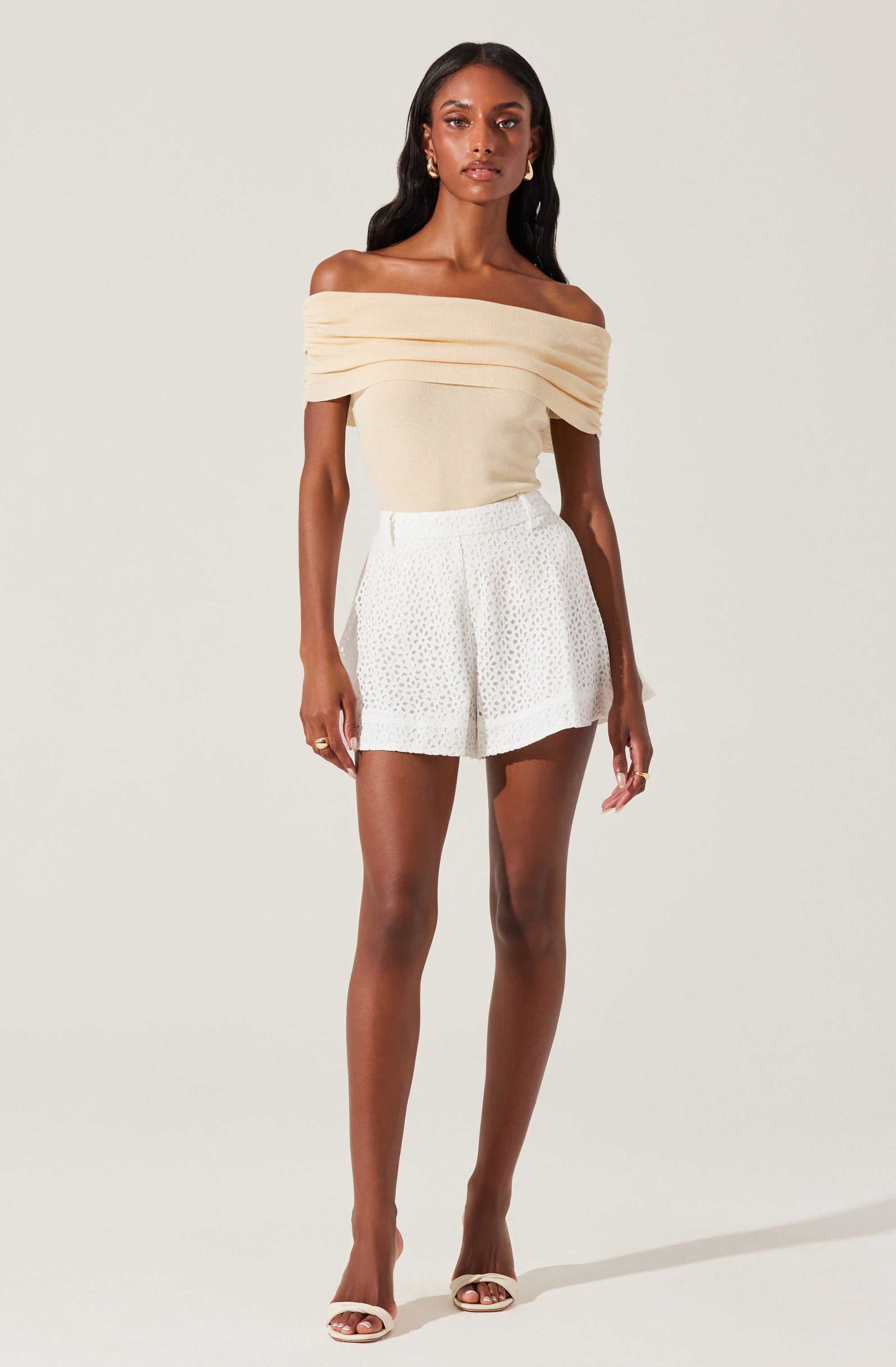A woman is wearing an off-the-shoulder, cream-colored top with draped, layered fabric around the neckline. She pairs it with high-waisted white shorts that have a subtle textured or dotted pattern. She completes her outfit with white open-toe heels.
