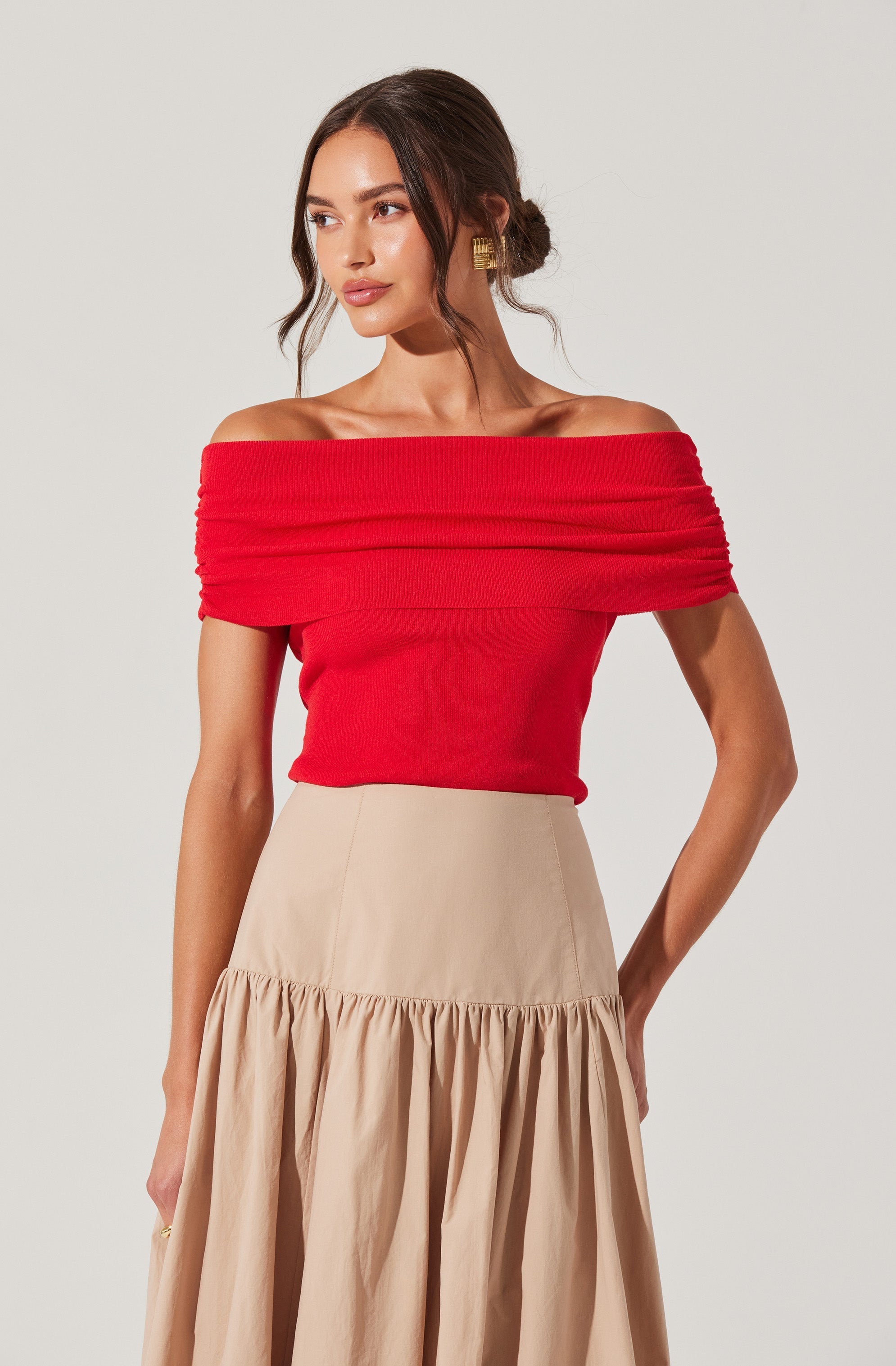 A woman is wearing an off-the-shoulder red top with horizontal ruching across the neckline and sleeves, paired with a high-waisted, beige skirt that has a gathered, ruffled hem.