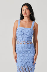 A woman is wearing a matching light blue crochet top and skirt set. The top has wide shoulder straps and a square neckline, featuring floral crochet patterns with some openwork sections at the midriff. The skirt is high-waisted and falls below the knee, also made of floral crochet patterns with openwork details.