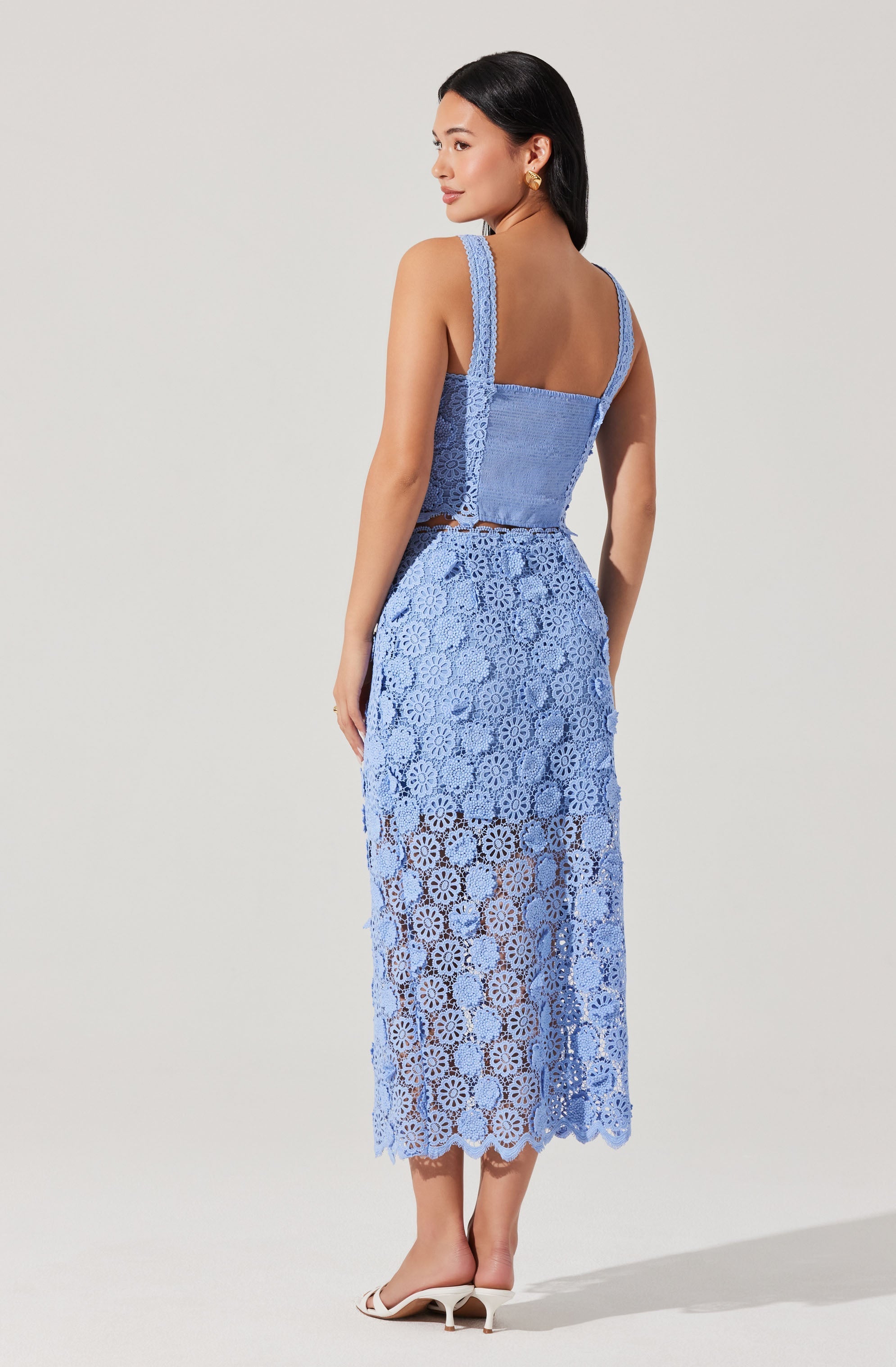 A woman is wearing a two-piece outfit made of blue lace. The top is a sleeveless, fitted tank with wide straps and a square neckline. The matching long skirt is also made of blue lace with a floral pattern, fitted at the waist and flowing down to her ankles with a scalloped hem. She is wearing white open-toe heels.