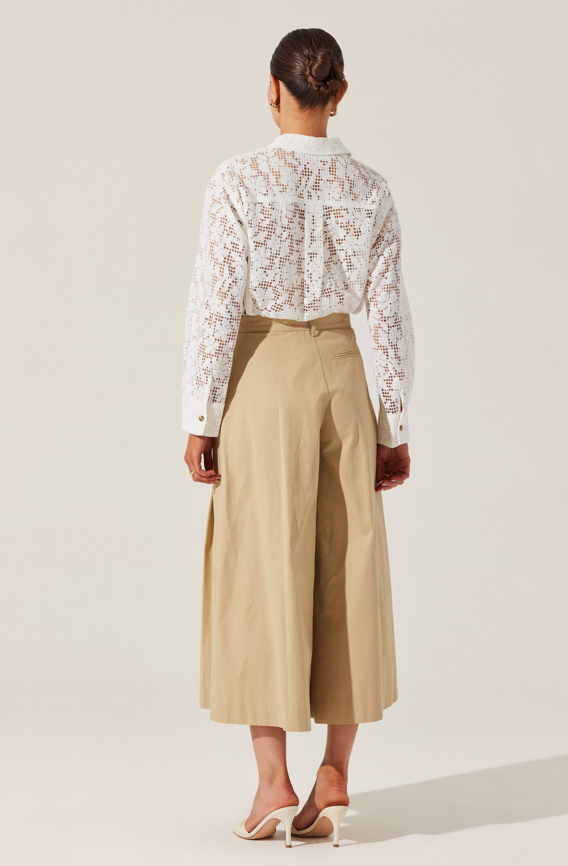 A woman is wearing a long-sleeved white blouse with a crochet or lace pattern, featuring a collar and buttoned cuffs. She pairs it with high-waisted, wide-leg beige trousers that reach mid-calf. She completes the outfit with white heeled shoes.