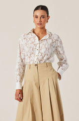 A woman is wearing a long-sleeved, white lace button-up shirt with a floral pattern and a classic collar. She has paired it with high-waisted, wide-leg beige trousers that feature pleats and a belt loop.