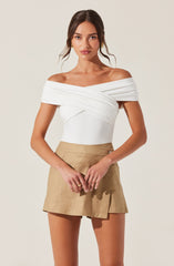 A woman is wearing an off-the-shoulder white top with draped and layered fabric details across the bust and shoulders. She is also wearing high-waisted beige shorts with a slight A-line shape, a front button closure, and side pockets.