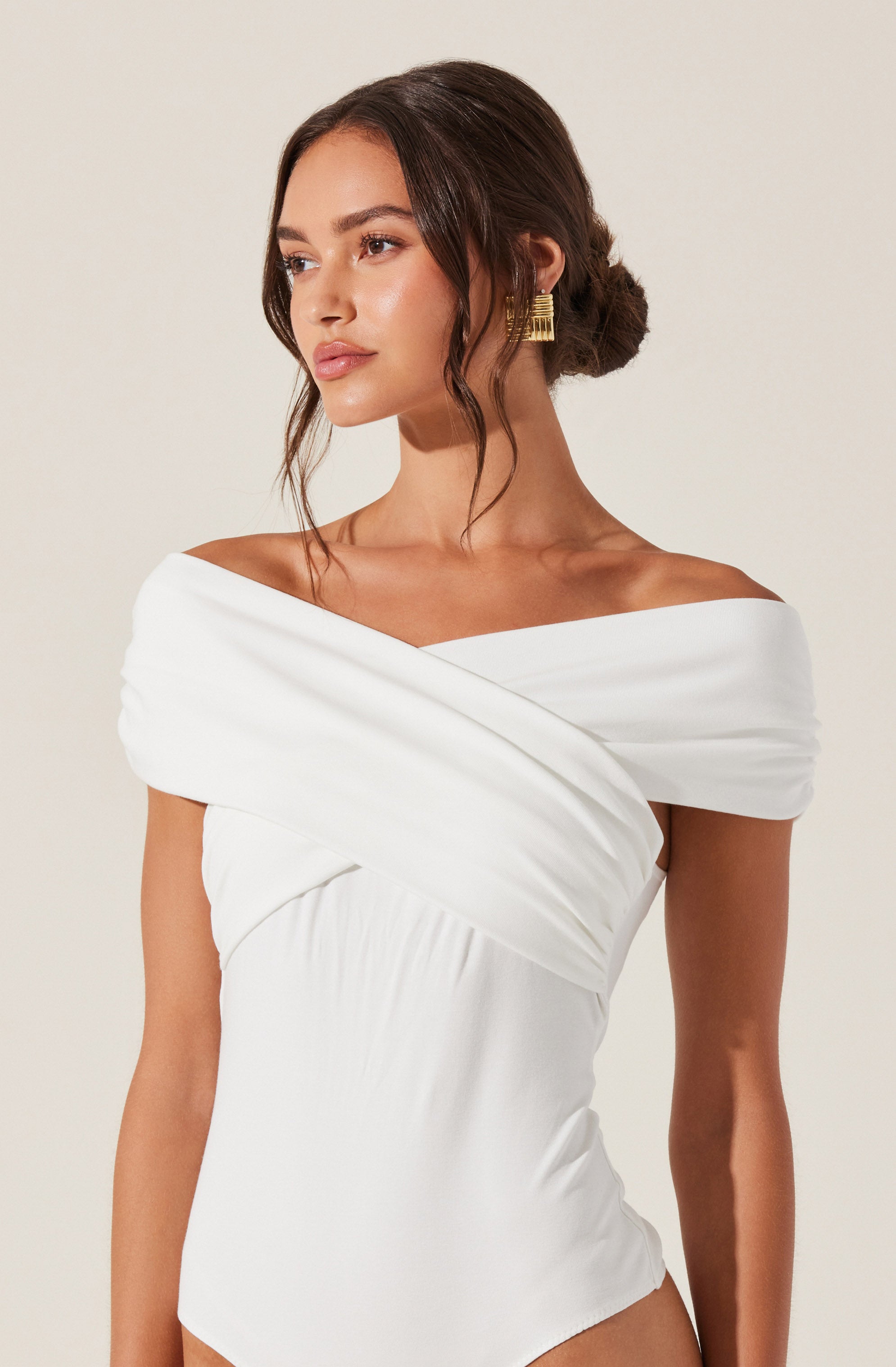 A woman is wearing an elegant white off-the-shoulder dress with wide, pleated fabric draped across the shoulders and bodice, creating a sophisticated look. She accessorizes with gold statement earrings and has her dark hair styled in a low, loose bun with soft waves framing her face.