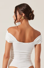 A woman is wearing an off-the-shoulder white top with ruched short sleeves. The top fits snugly and is made of a smooth, slightly shiny fabric. She has her dark brown hair styled in a low, twisted bun and is accessorized with large, gold, rectangular earrings.