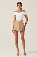 A woman is wearing an off-the-shoulder white top with draped, twisted fabric across the chest, paired with a beige wrap-style mini skirt that has a side button detail. She is also wearing white heeled sandals.