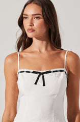 A woman is wearing a white sleeveless top with thin shoulder straps. The top features black ribbon details along the neckline with a small black bow in the center. The fabric appears lightweight and comfortable.
