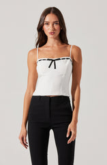 A woman is wearing a white sleeveless top with thin spaghetti straps. The top features black trim and a small black bow at the center of the neckline. She is also wearing high-waisted black pants.