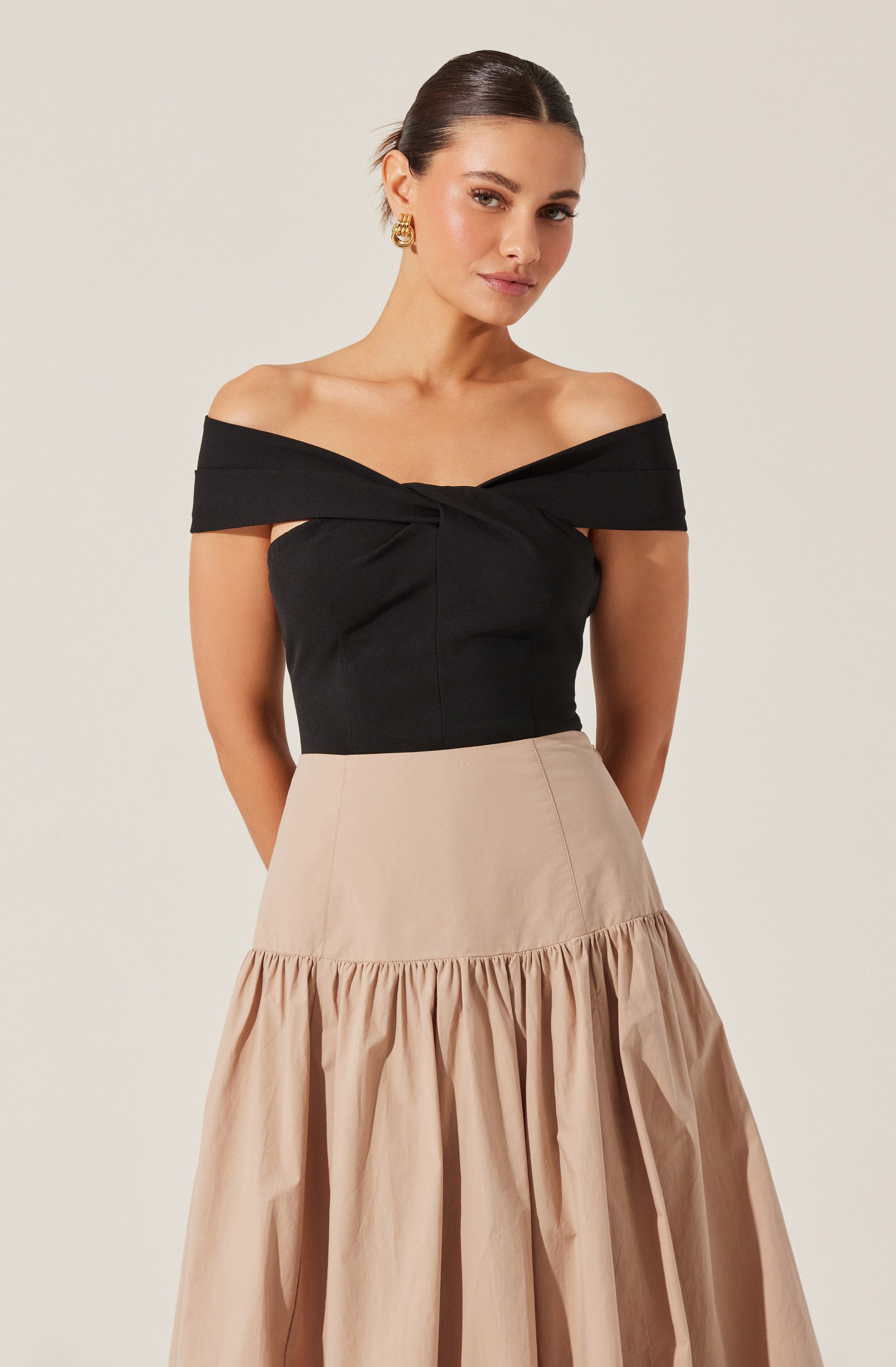 A woman is wearing an off-the-shoulder black top with a twisted knot detail at the front. She has paired it with a beige, high-waisted, A-line skirt that features a gathered ruffle at the hem. She accessorizes with gold hoop earrings.