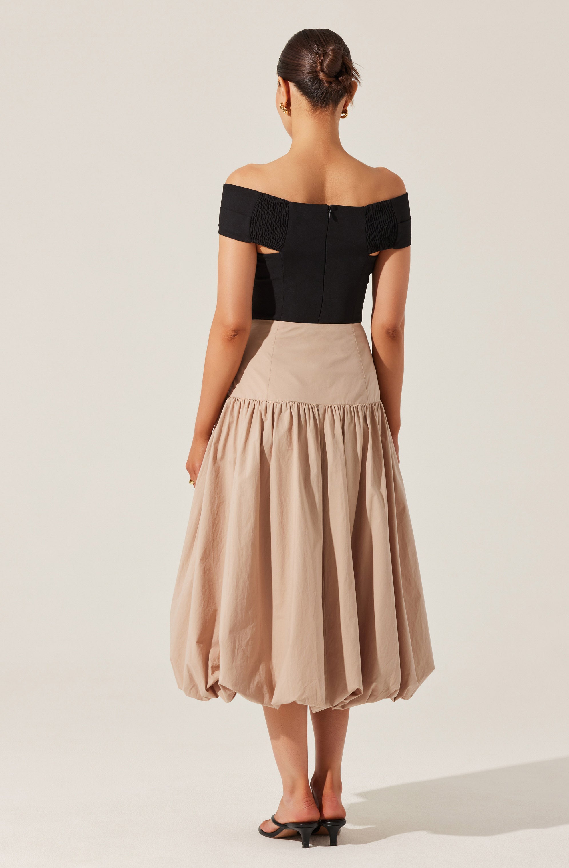 The woman is wearing an off-the-shoulder black top with textured band details on the sleeves, paired with a high-waisted, beige, gathered midi skirt that has a voluminous, bubble-like hem. She is also wearing black heeled sandals.