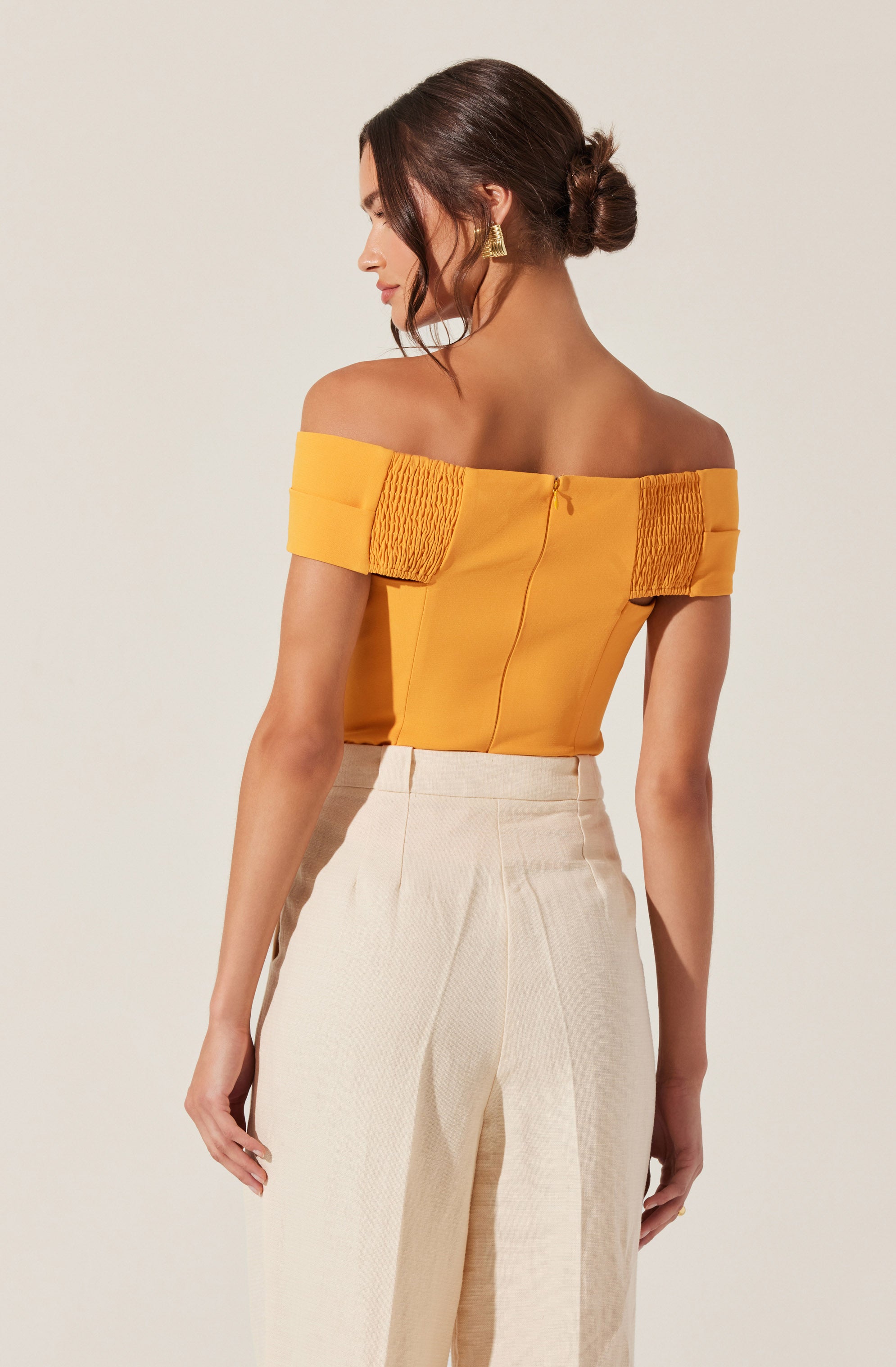A woman is wearing an off-the-shoulder, mustard yellow top with gathered fabric details on the sleeves and a zipper closure in the back. She pairs it with high-waisted, wide-leg cream-colored trousers. She has dark brown hair styled in a low bun and is accessorized with large, gold triangular earrings.