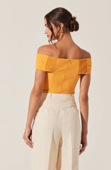 A woman is wearing an off-the-shoulder, mustard yellow top with gathered fabric details on the sleeves and a zipper closure in the back. She pairs it with high-waisted, wide-leg cream-colored trousers. She has dark brown hair styled in a low bun and is accessorized with large, gold triangular earrings.