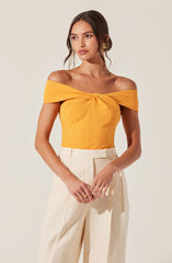 A woman is wearing an off-the-shoulder yellow top with a twisted knot detail at the front. She pairs it with high-waisted, wide-leg cream trousers. She has long, dark hair styled in loose waves and is accessorized with gold earrings.