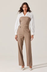 A woman is wearing a white, long-sleeved, collared shirt with vertical stripes. Over the shirt, she has on a strapless, beige tailored jumpsuit that fits snugly at the top and flows into wide-leg trousers. She is also wearing pointed white heels.