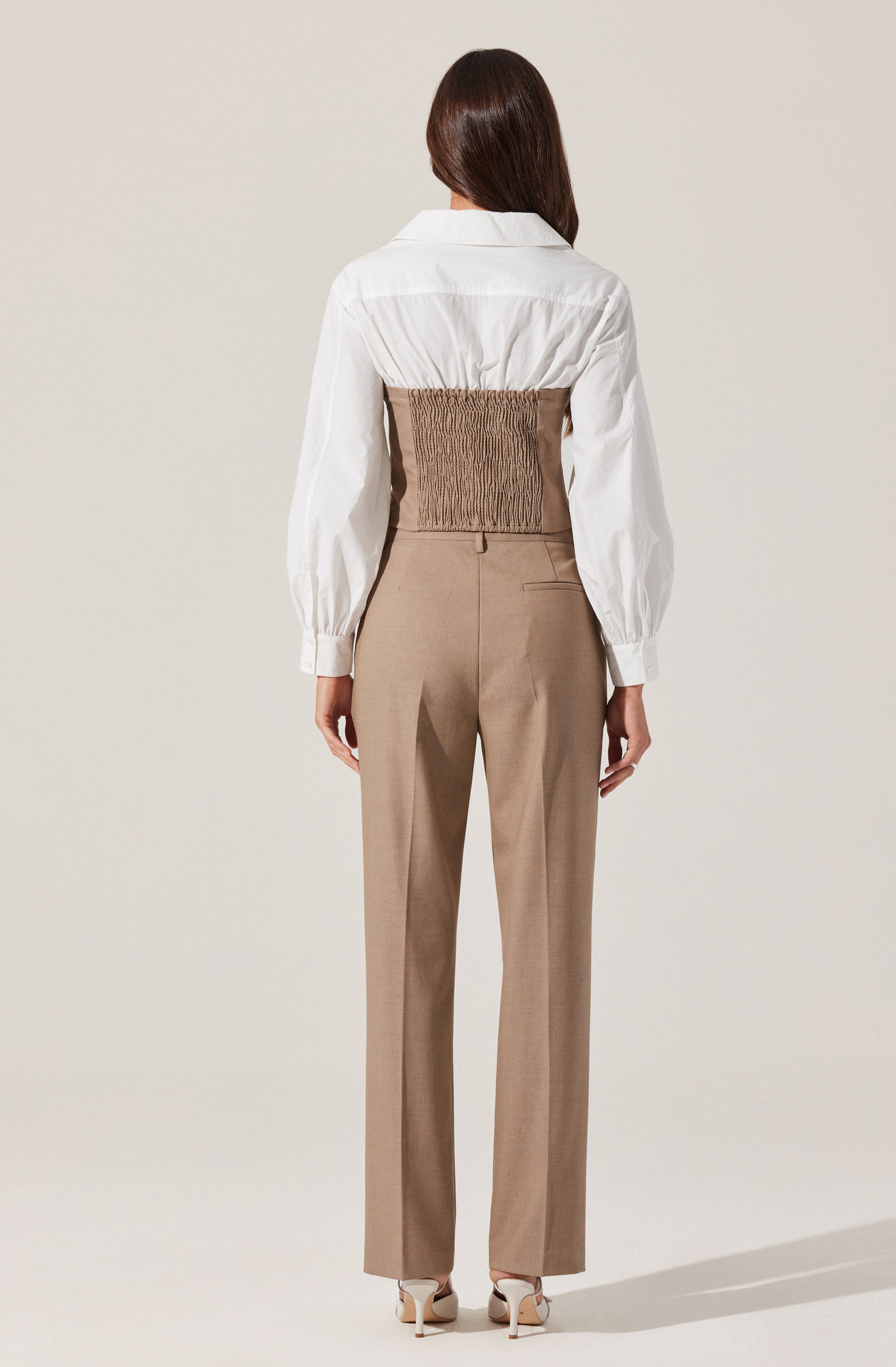 A woman is wearing a white long-sleeve shirt with a loose fit and a collar. The shirt has gathered cuffs and is tucked into high-waisted beige trousers. The trousers are tailored, straight-leg, with a back pocket visible on the right side. She is also wearing beige high-heeled shoes.