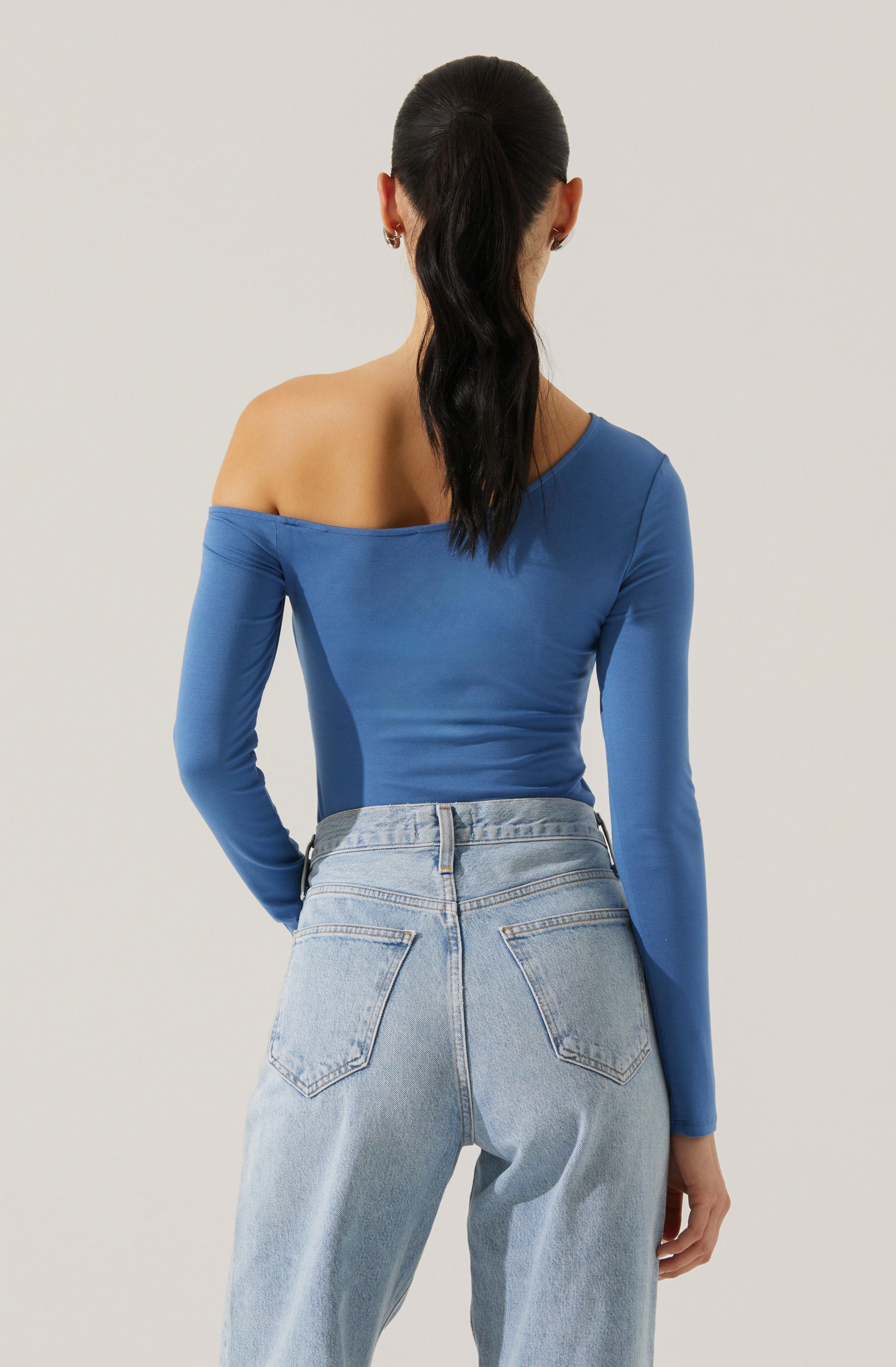 A woman is wearing a fitted, off-the-shoulder, long-sleeve top in a bright blue color. She pairs it with high-waisted, light-wash denim jeans.