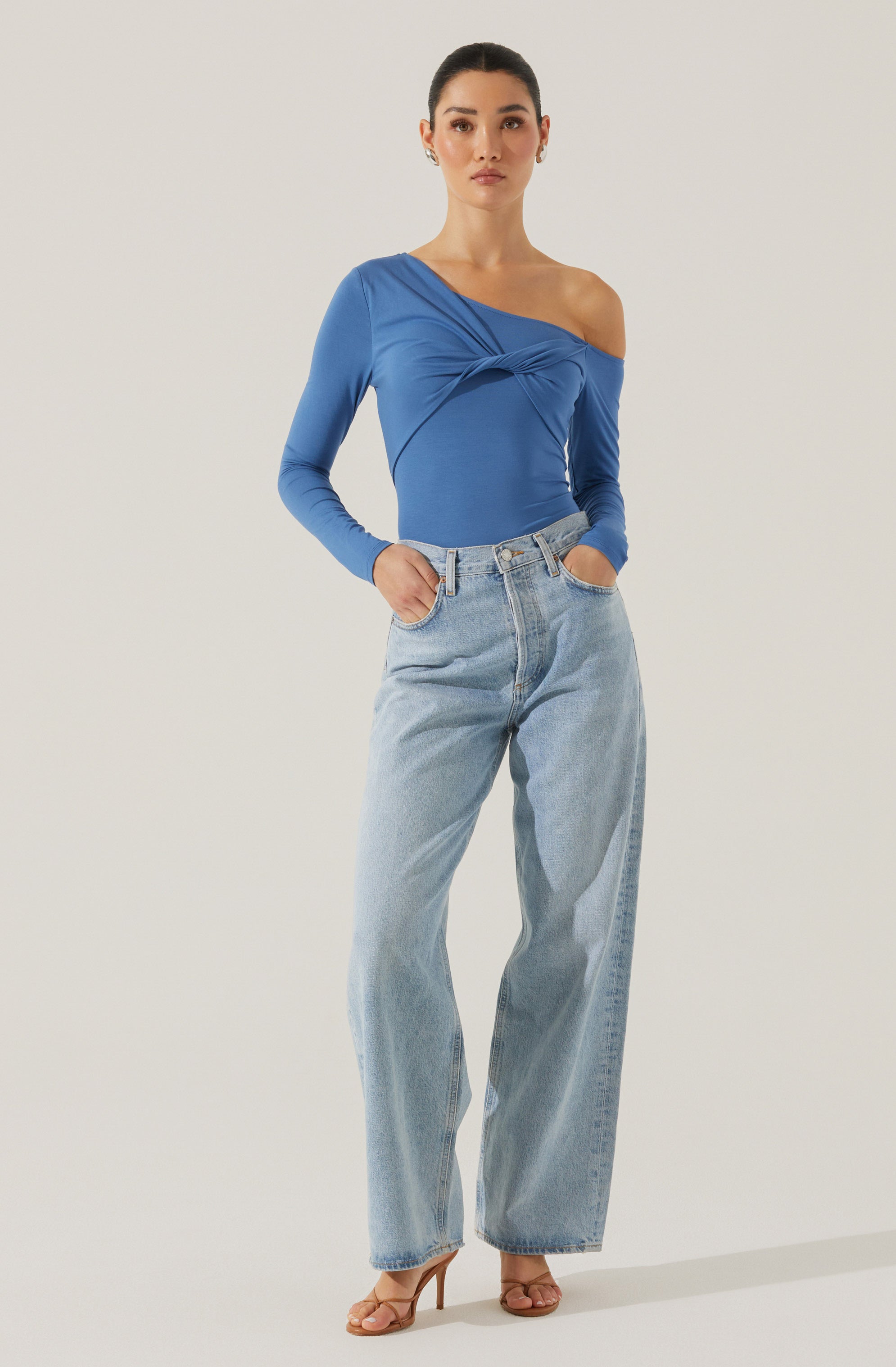 A woman is wearing a blue, asymmetrical, long-sleeved top with a twisted knot detail across the chest and an off-the-shoulder neckline on the right side. She pairs the top with high-waisted, light-wash, wide-leg jeans. She is also wearing nude-colored high heels.