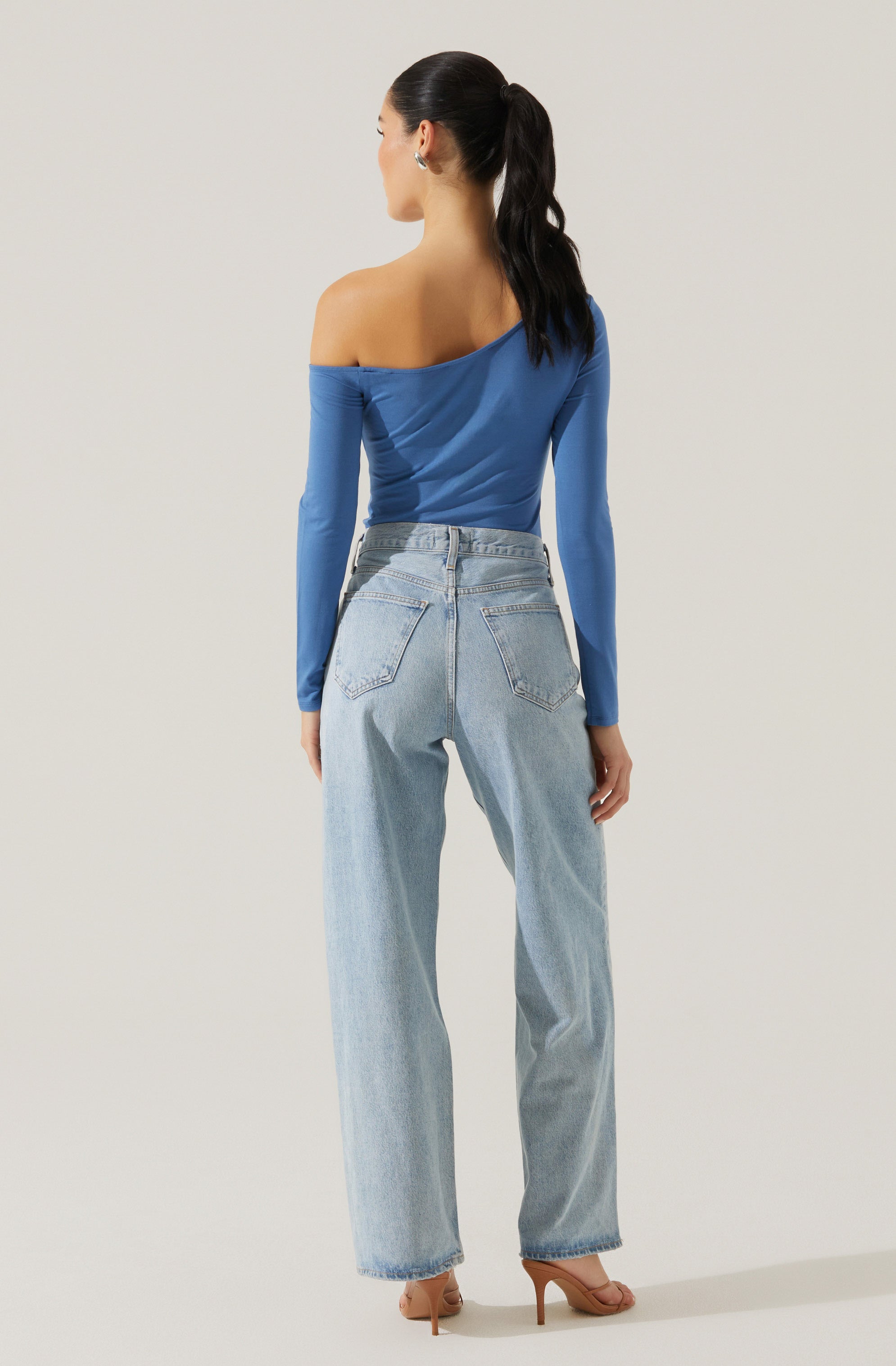 A woman is wearing a fitted, long-sleeve, off-the-shoulder blue top with a ruched detail on the side. She pairs it with high-waisted, light-wash, straight-leg jeans that have a classic five-pocket design and slightly frayed hems. She completes the outfit with nude high-heeled sandals.